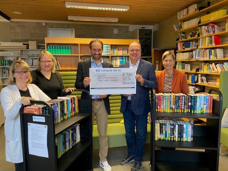 Personen hinter mobilen Leseregalen lächeln in die Kamera und halten einen Scheck hoch